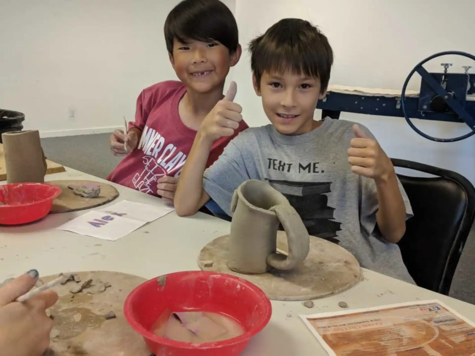 a couple of kids sitting at a table with a clay pot and a bowl of food