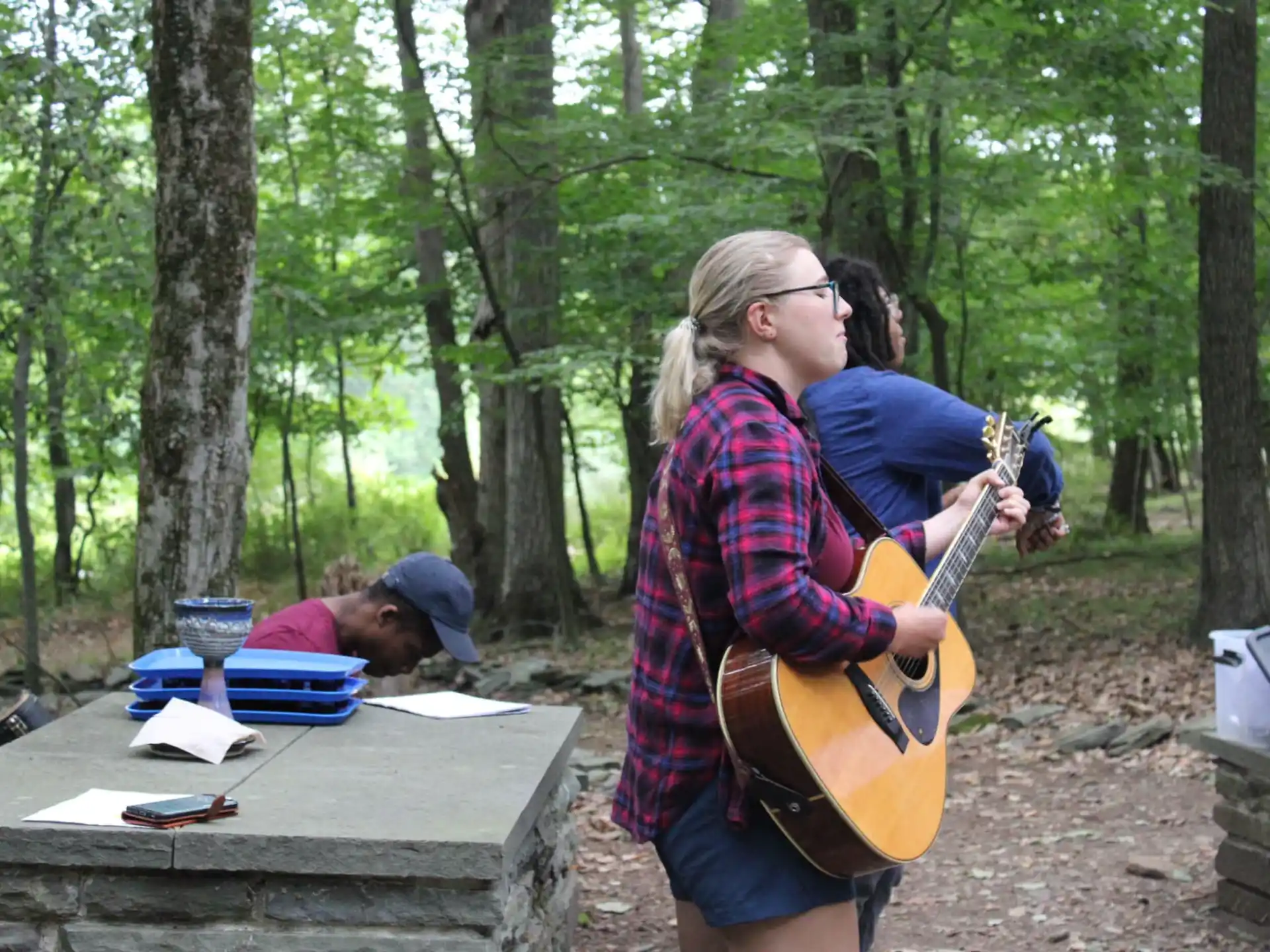 a woman playing a guitar next to a man sitting at a picnic table