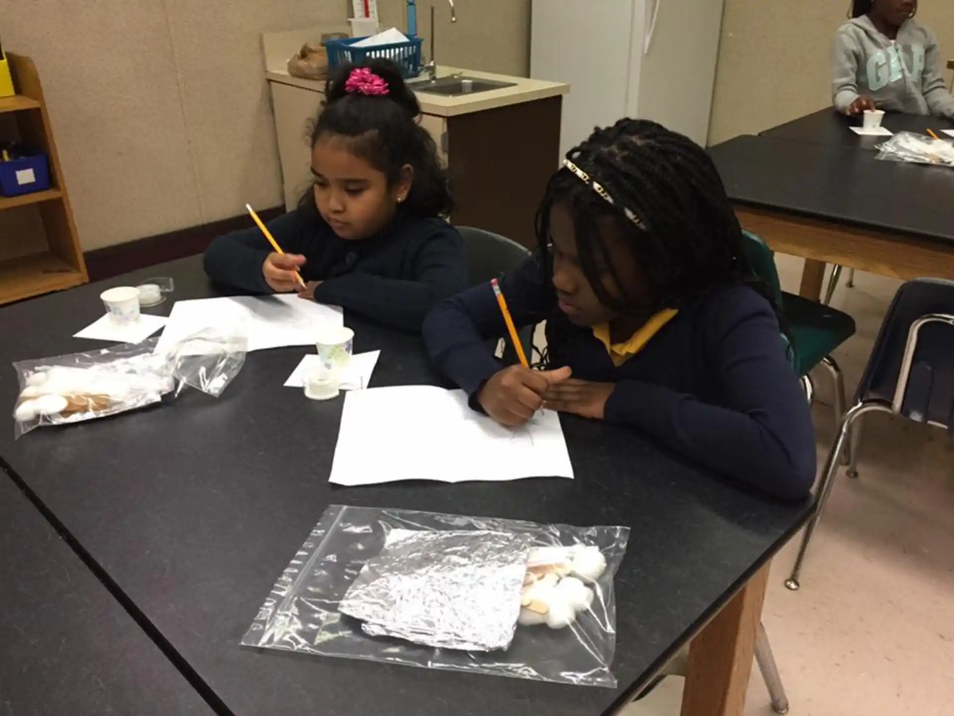 a couple of girls sitting at a table with a black board and pencils
