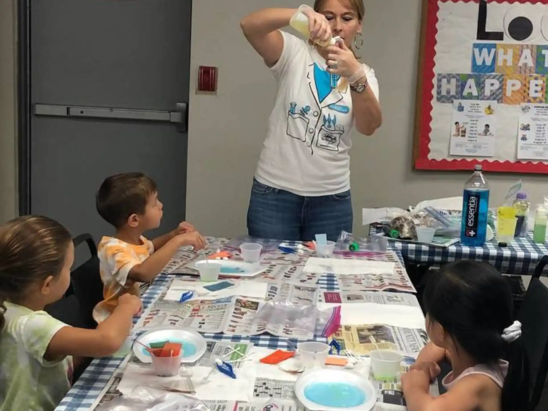 a woman standing in front of a table with children sitting at it