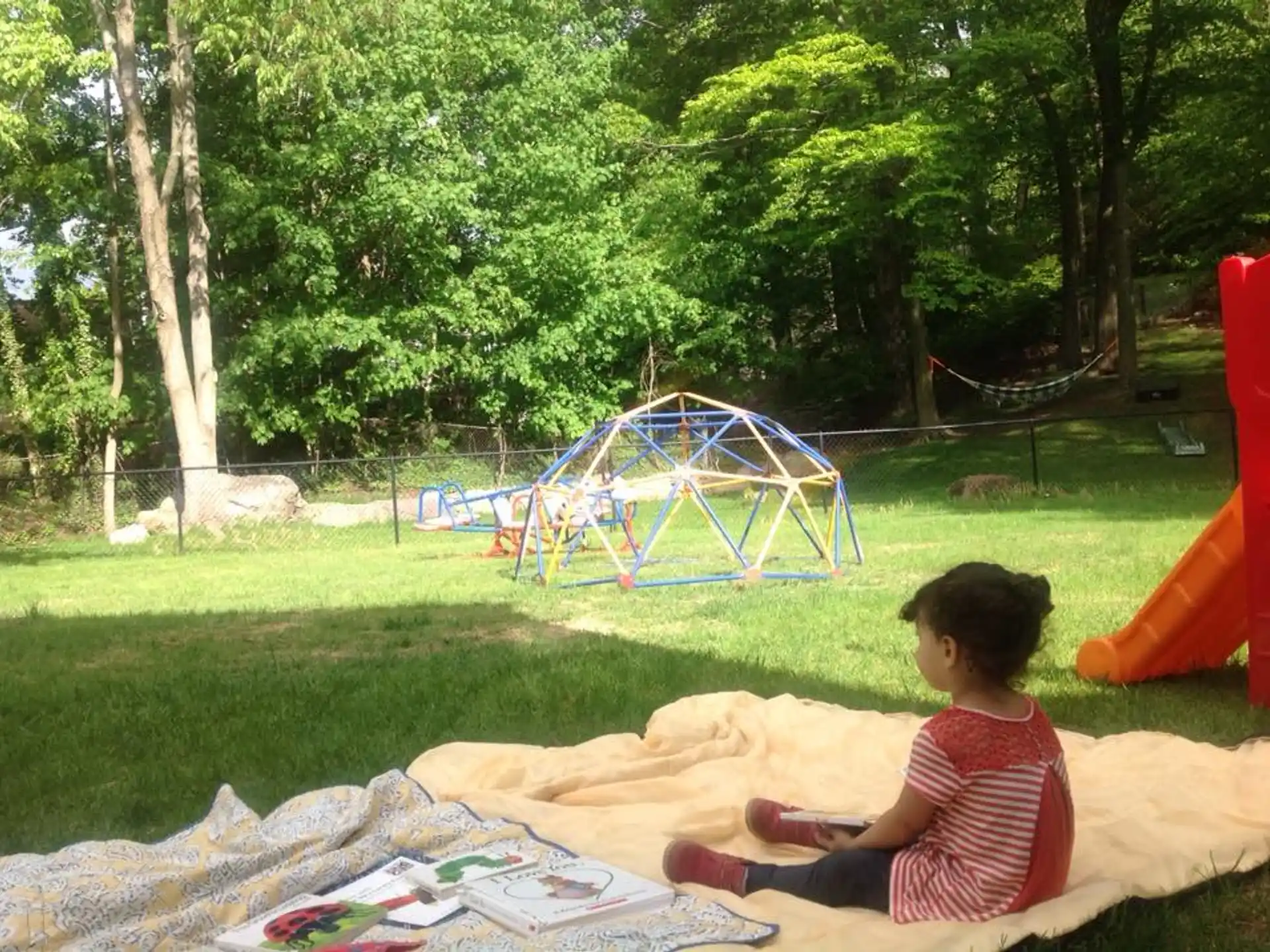 a child sitting on a blanket in a grassy area with a trampoline and trees in the