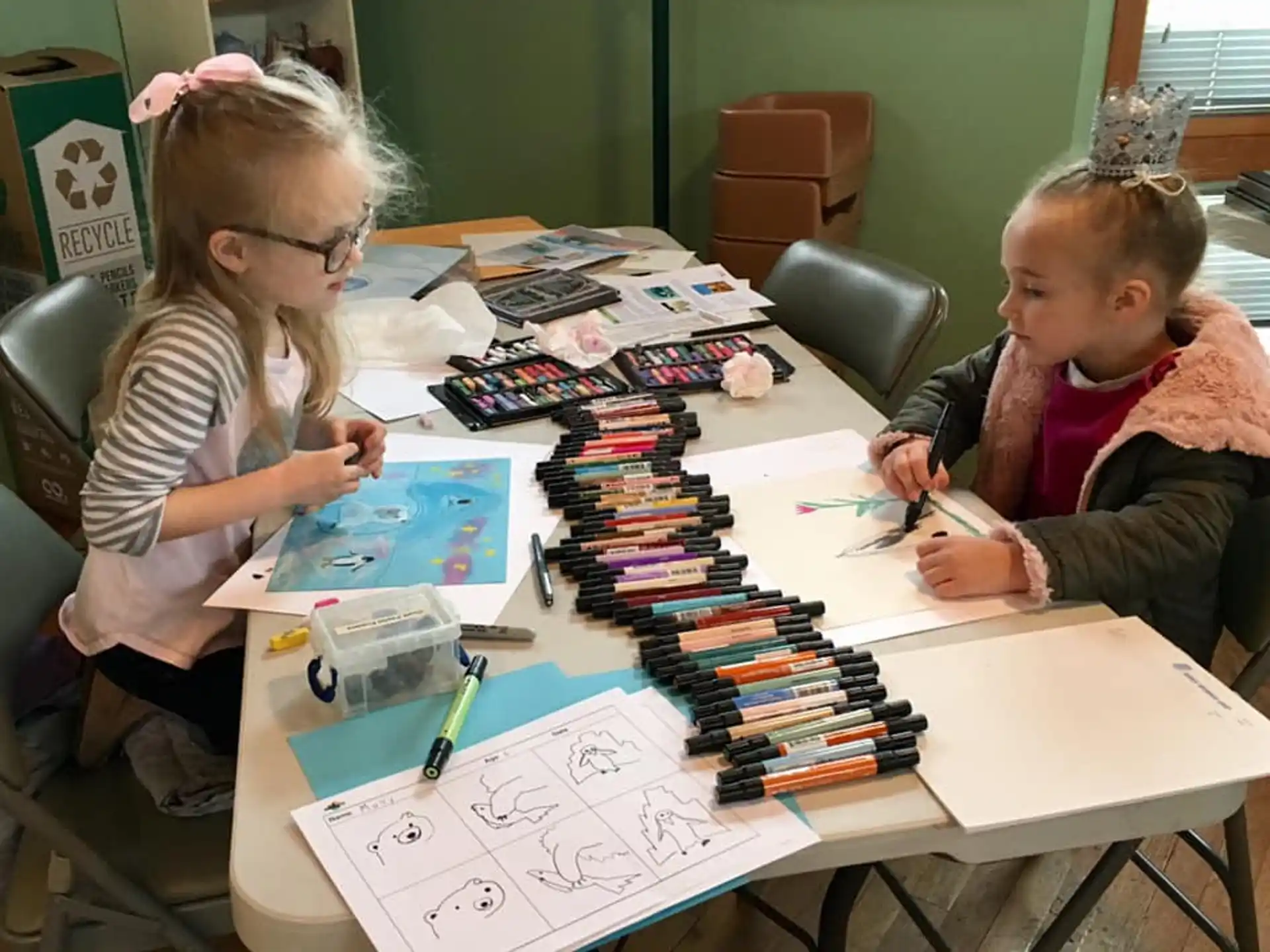 a couple of young girls sitting at a table with papers and pens