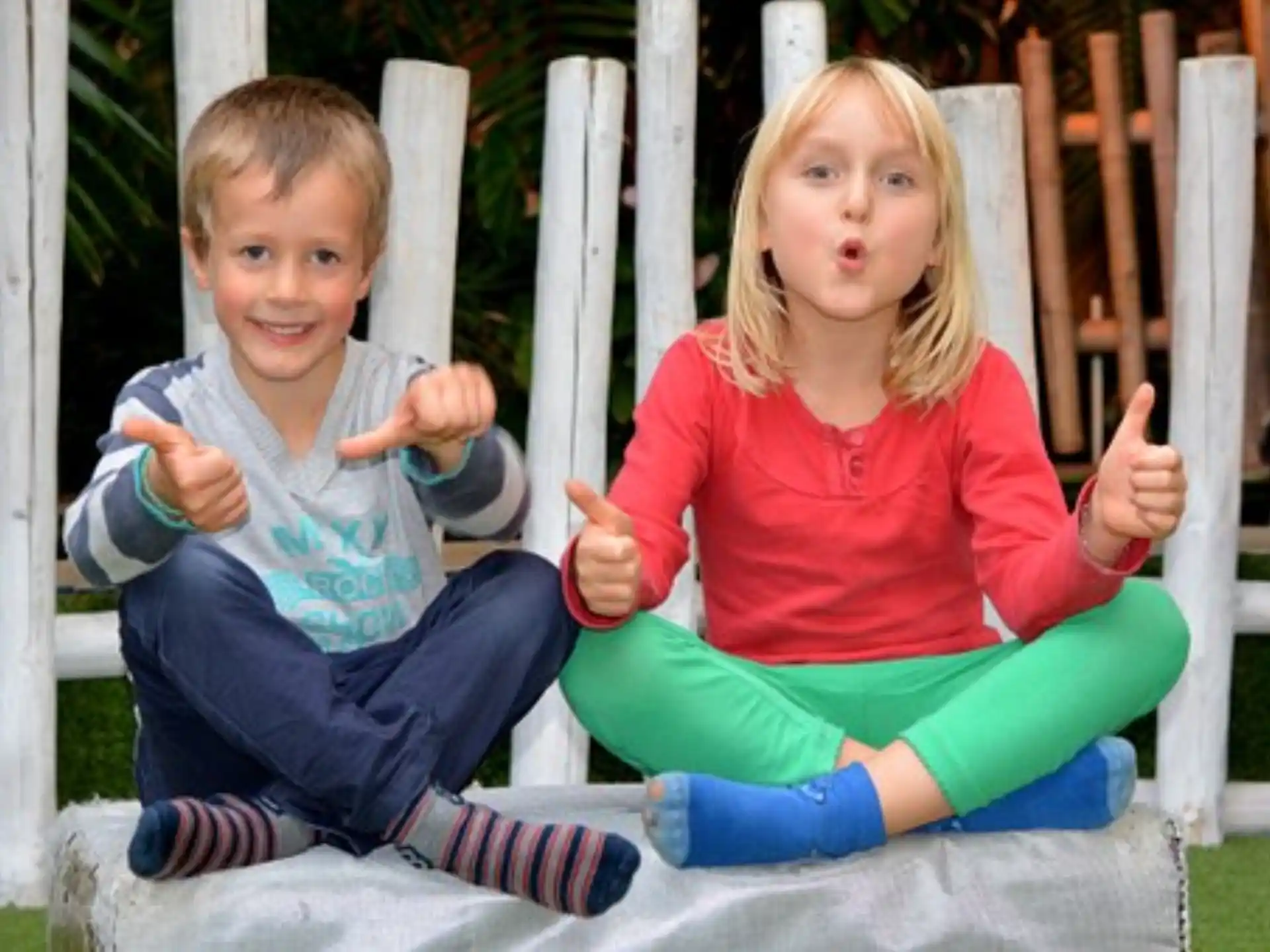 a boy and girl sitting on a bench and pointing at the camera
