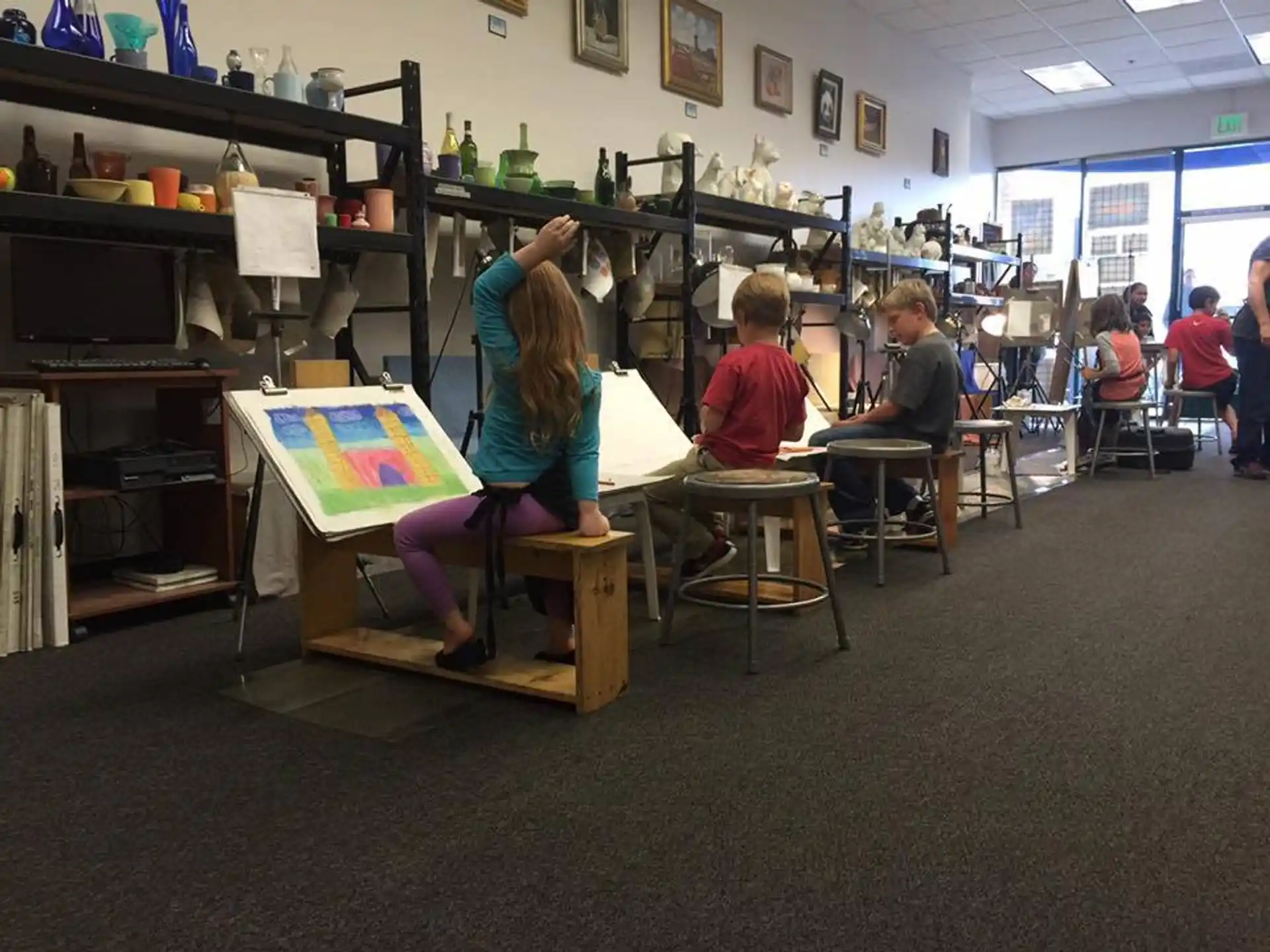 a group of people sitting at desks in a room with shelves and a wall with pictures on
