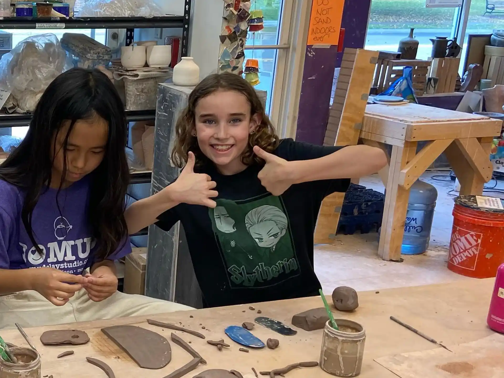a couple of girls sitting at a table with clay pots and a wood carving
