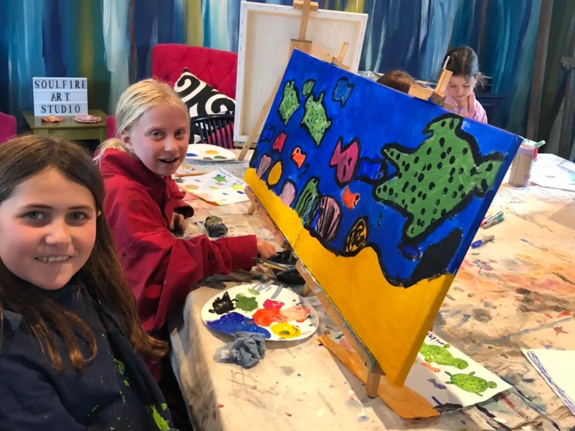 a couple of girls sitting at a table with a painting on it