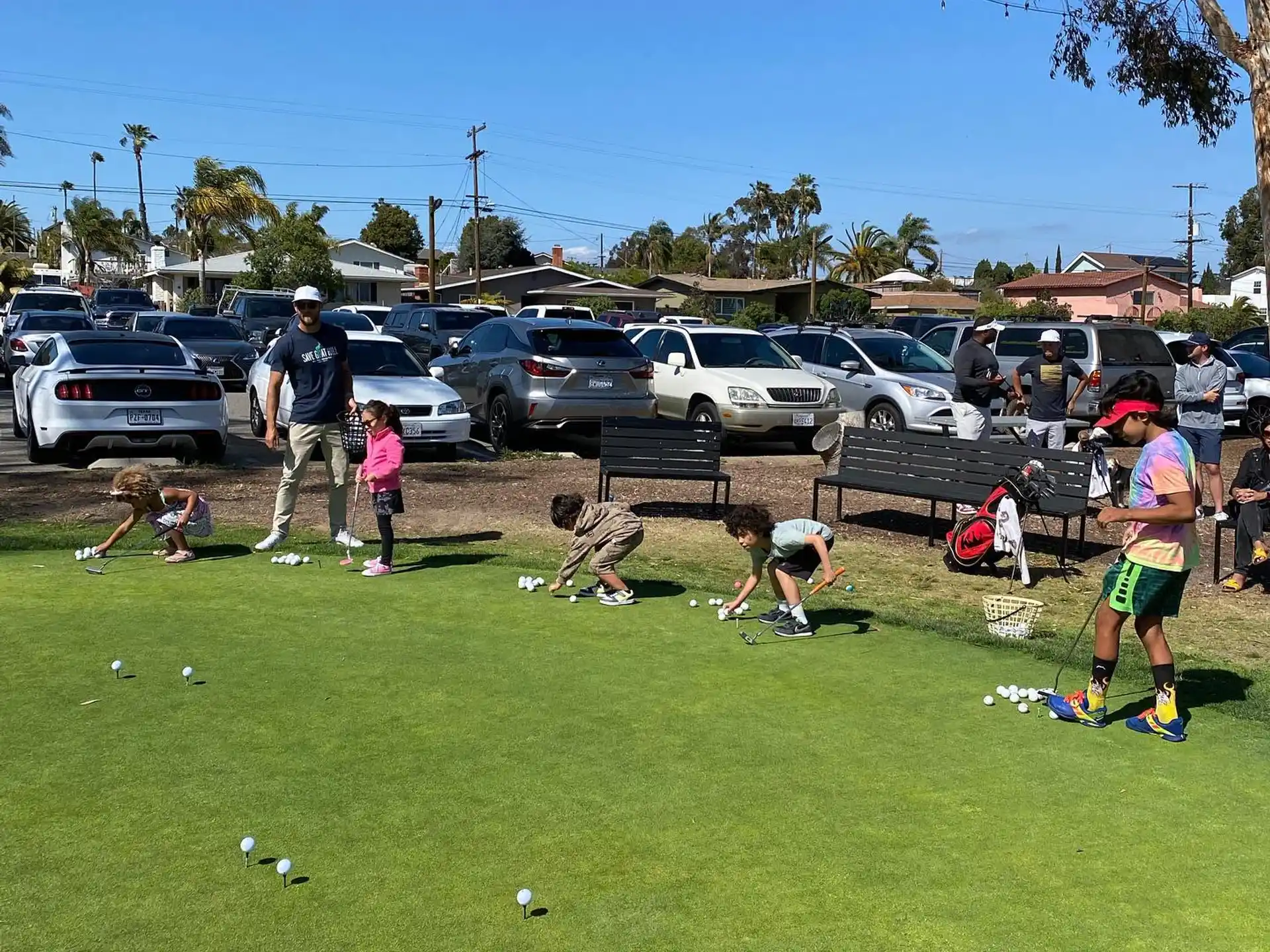 a group of people playing in a grassy field with cars and a parking lot in the background