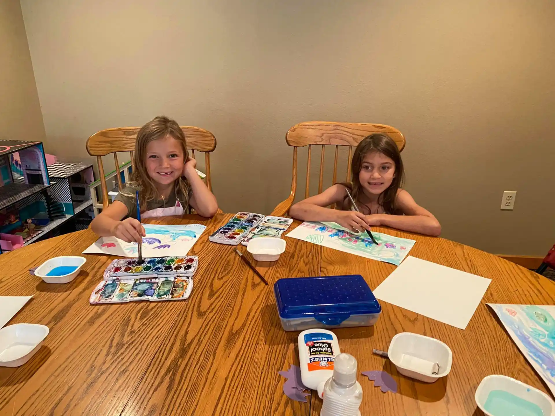 a couple of girls sitting at a table with books and drawings