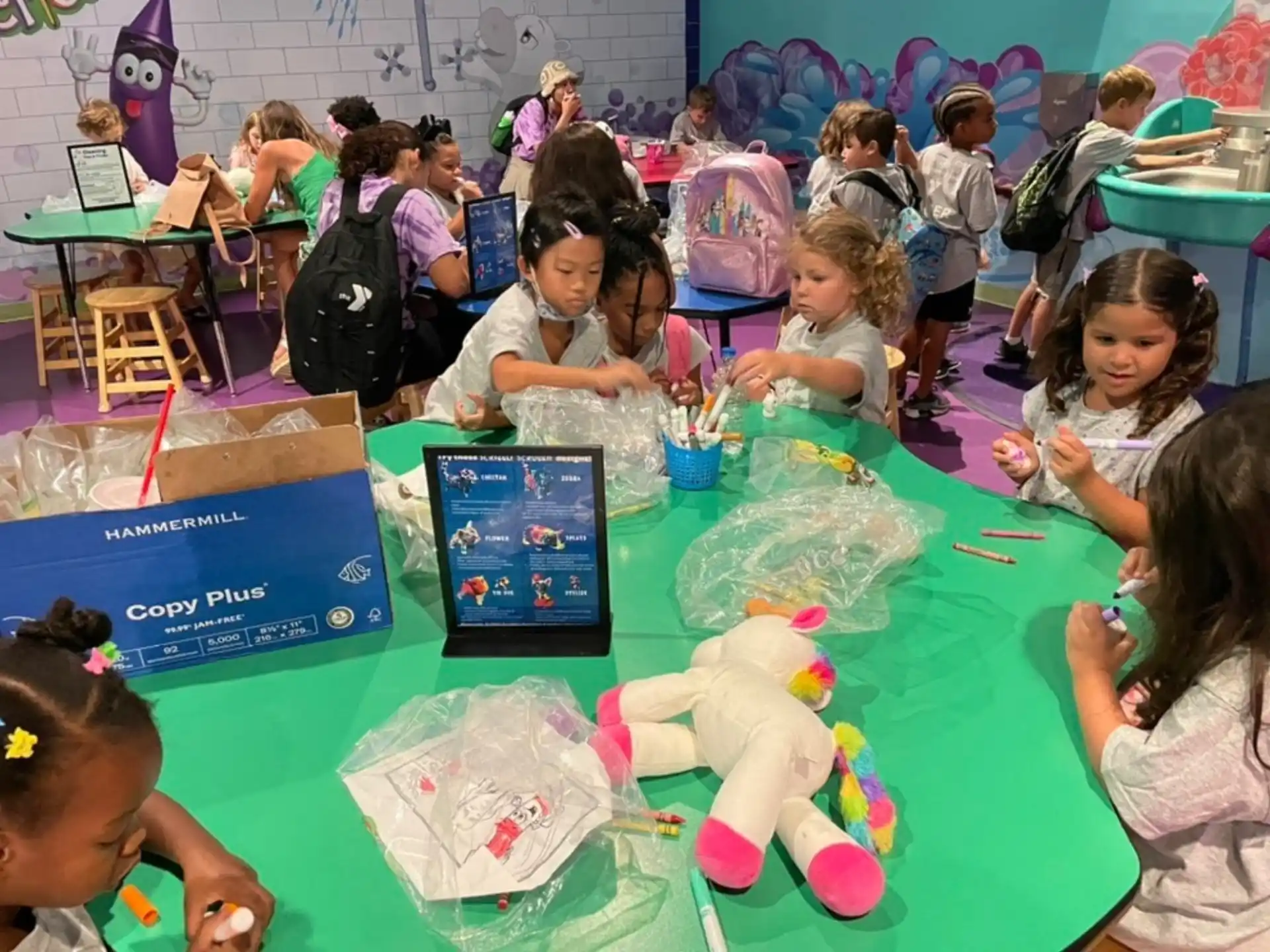 a group of children sitting at a table with a tablet and a stuffed animal