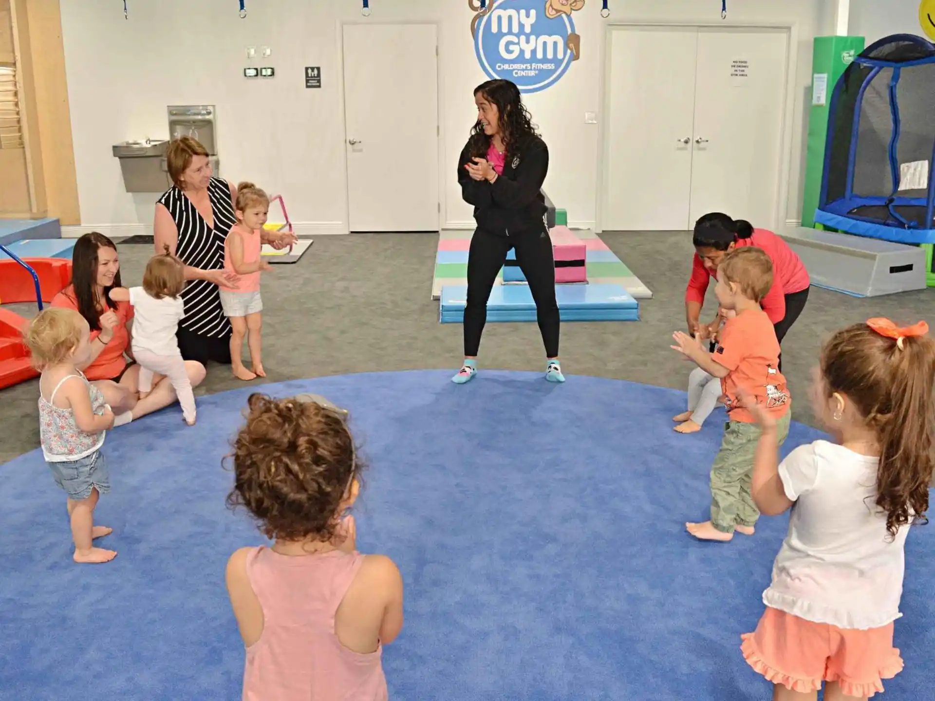 a group of children playing on a mat in a room with a person standing and a girl sitting