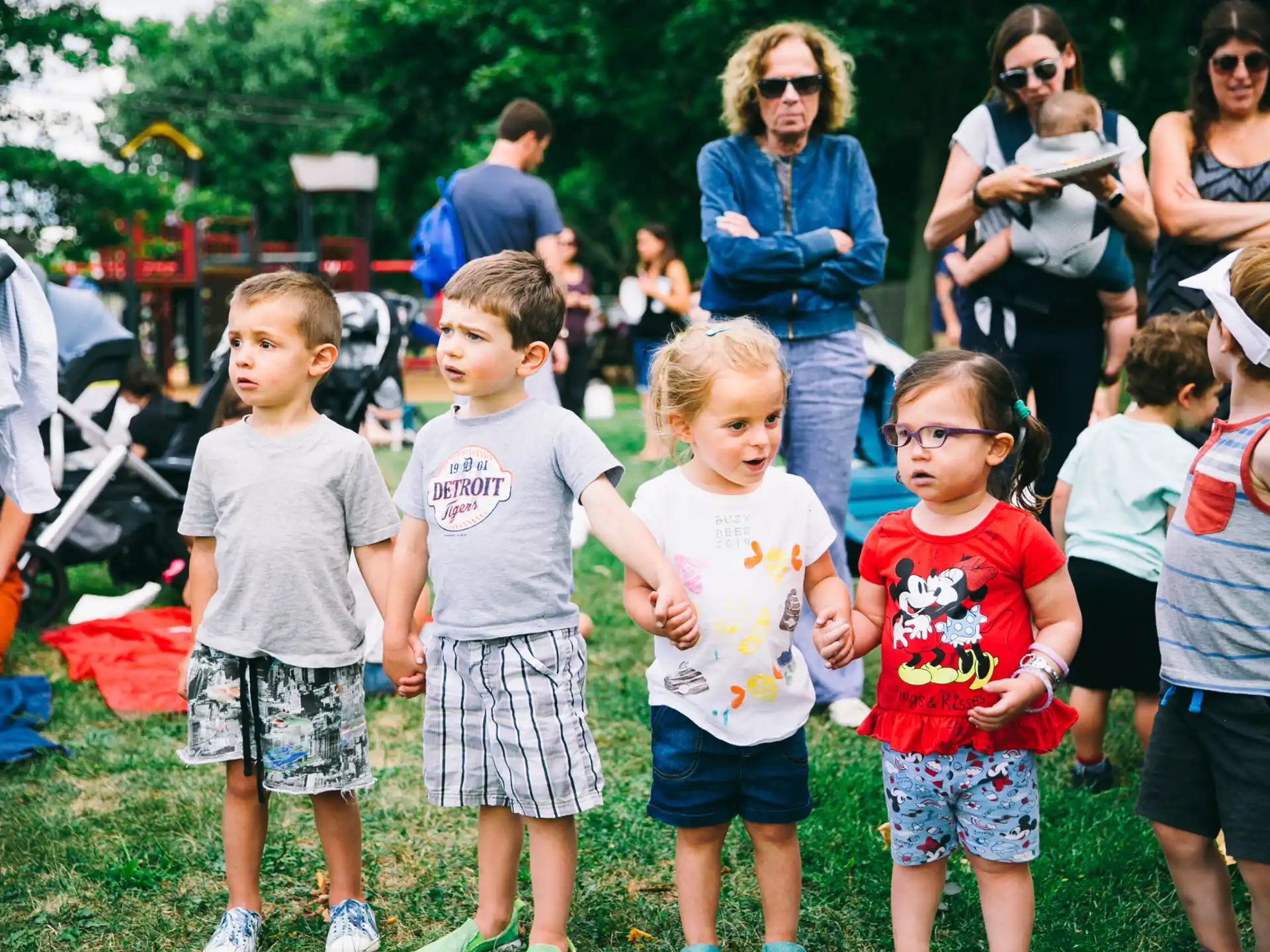 a group of children standing in a grassy area with people in the back