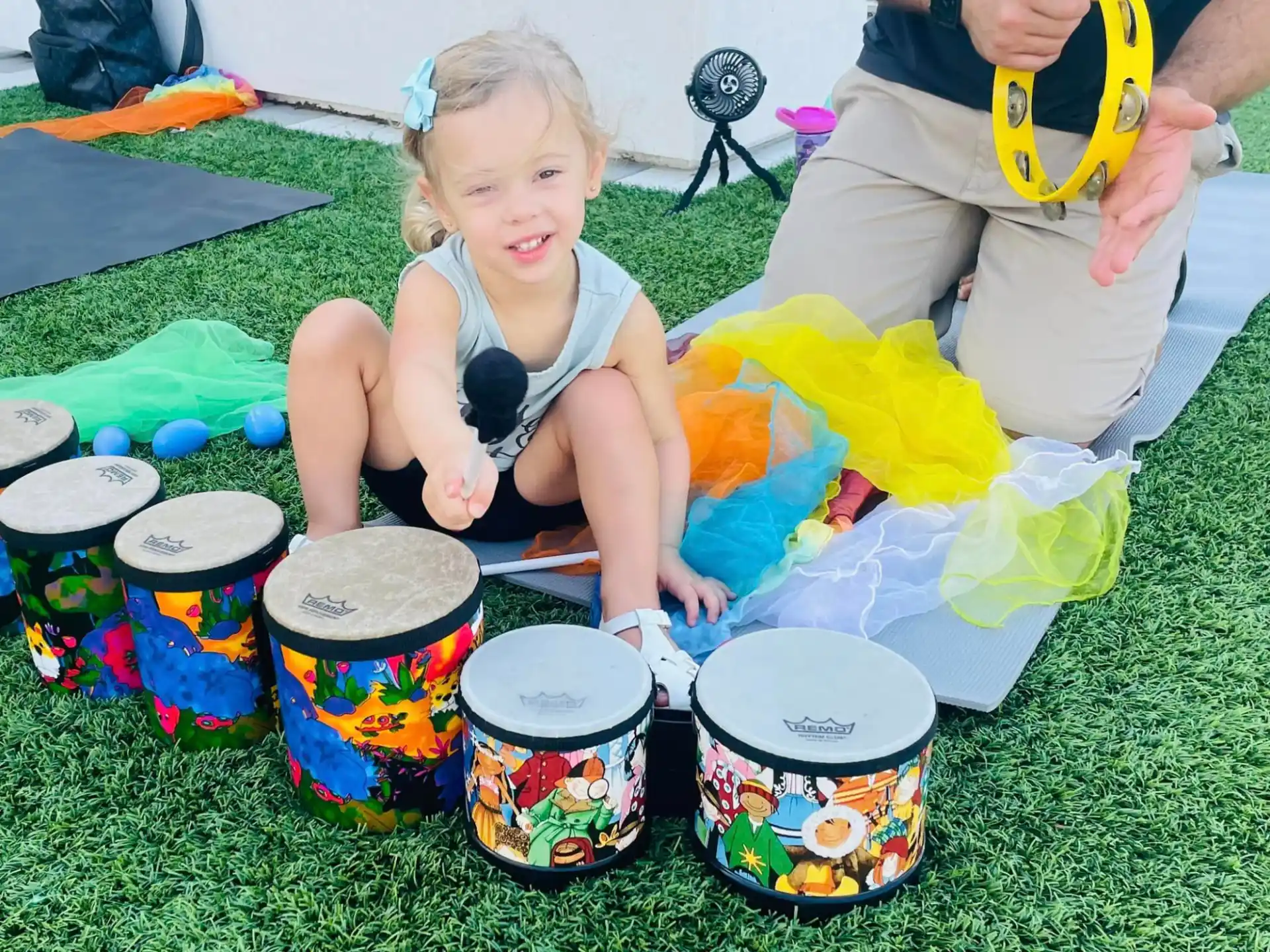 a girl sitting on the grass with drums and a person standing by her
