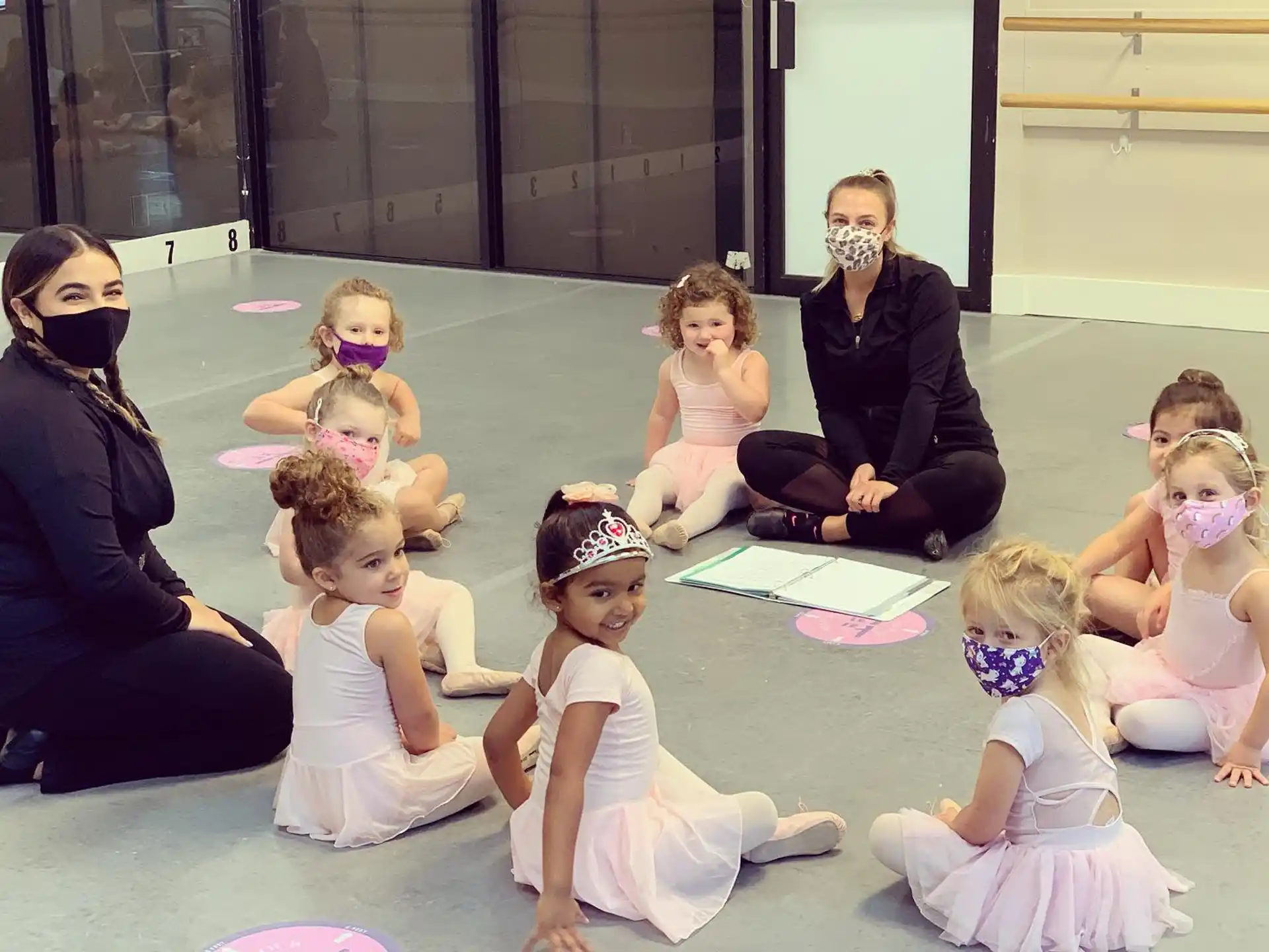 a group of children sitting on the floor with a person in a black dress and a white mask