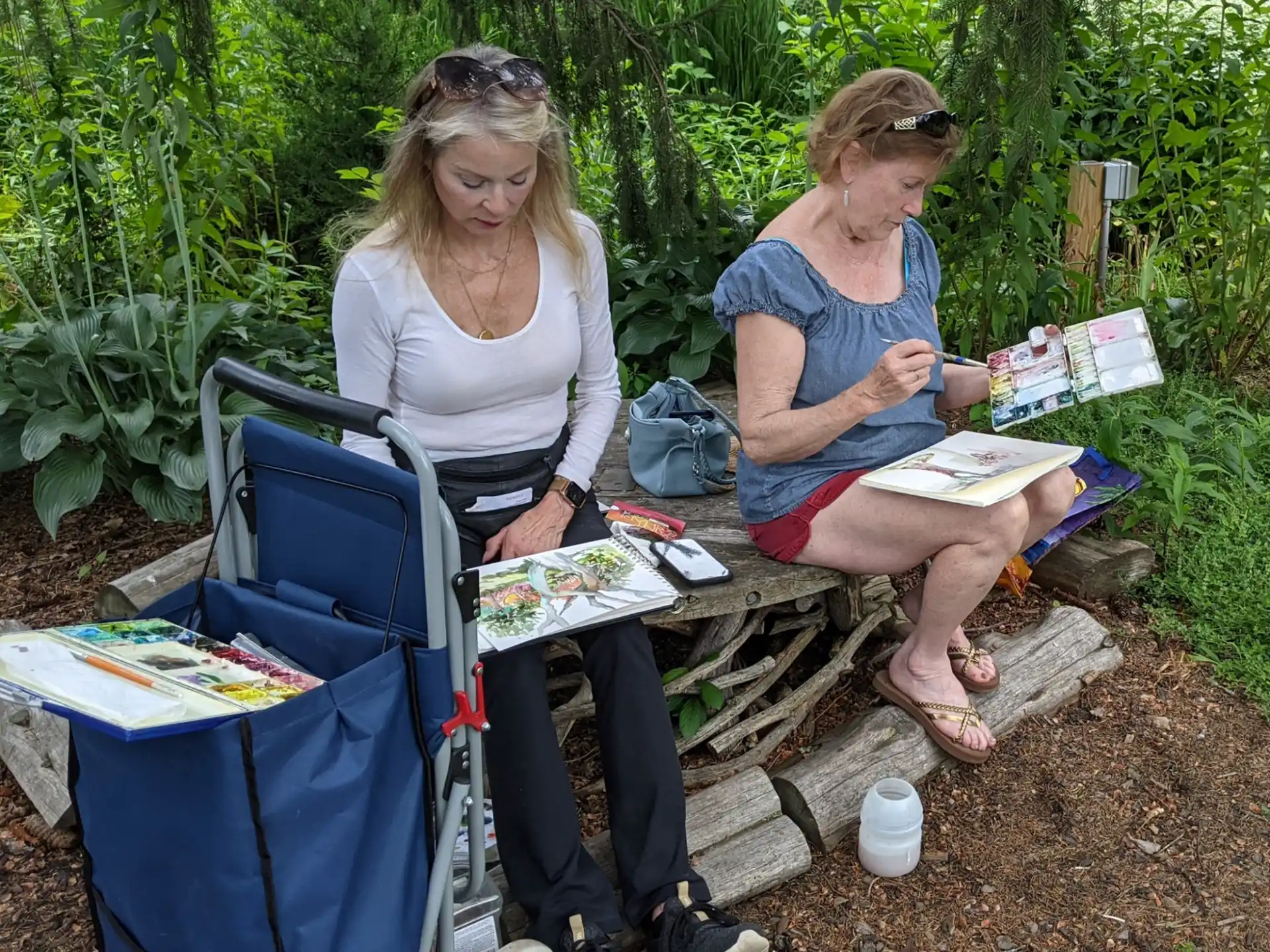 a couple of women sitting on a bench with books and a blue cart