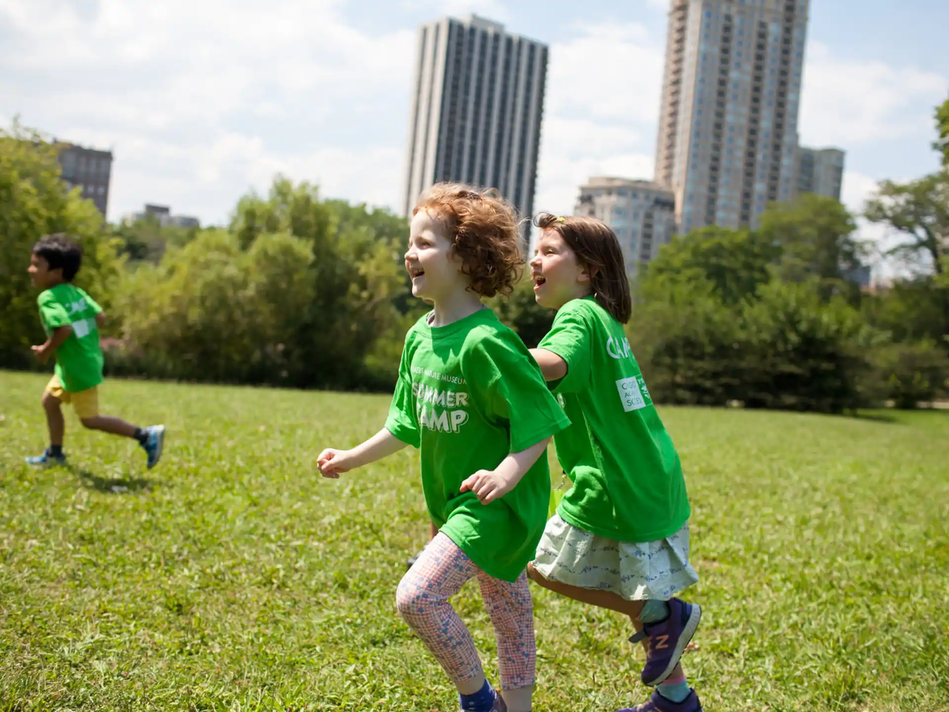 a group of kids running on a grass field with trees and buildings in the background