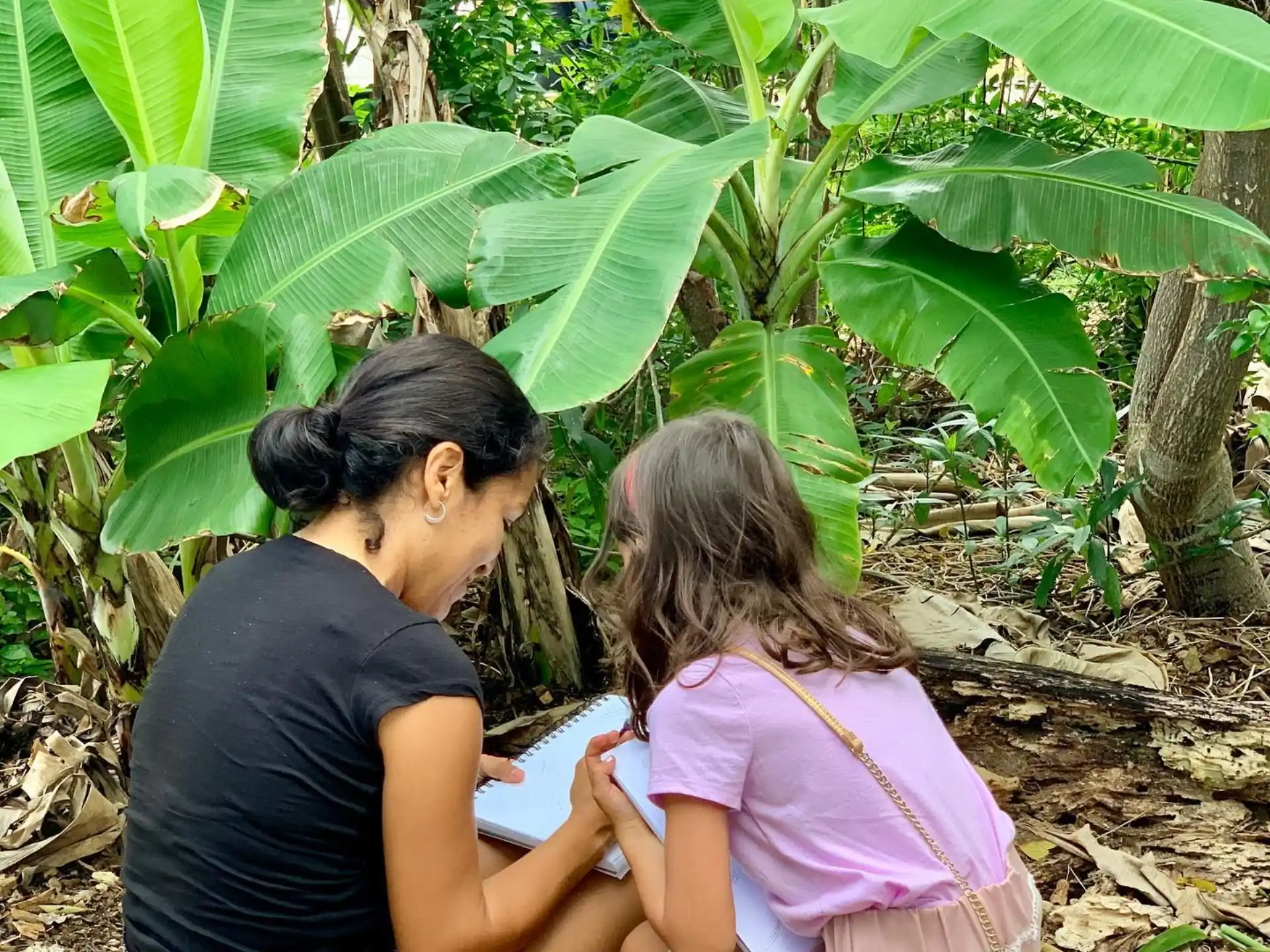 a person and a child sitting on the ground looking at a plant