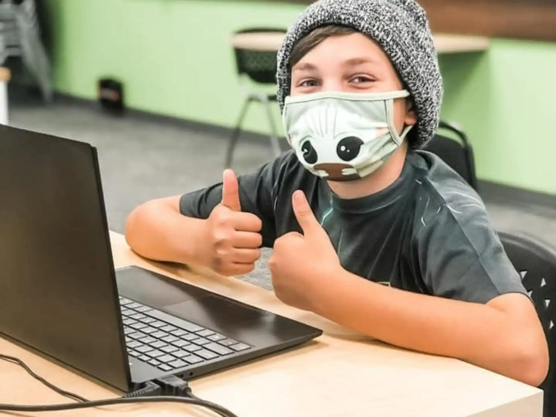 a person wearing a mask and sitting at a table with a laptop