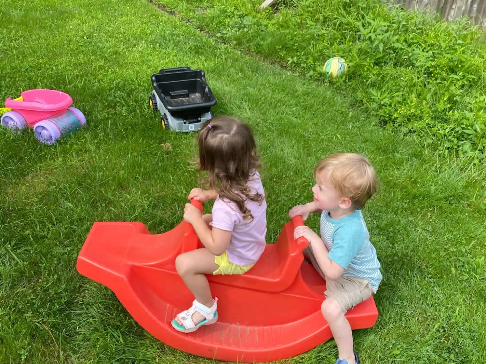 two children sitting on a red plastic toy in a grassy area
