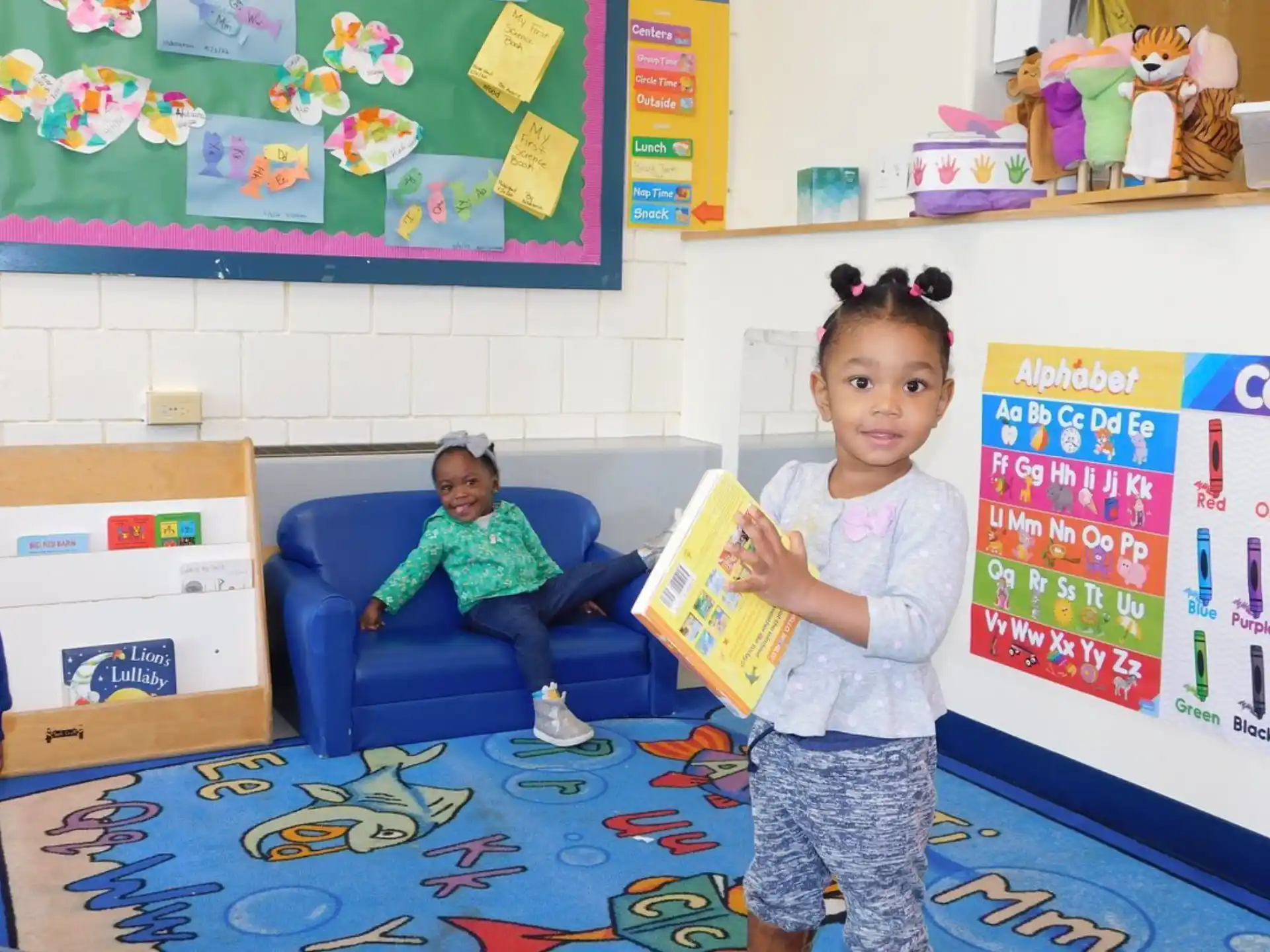 a young girl holding a book and a young girl sitting on a blue chair in a classroom