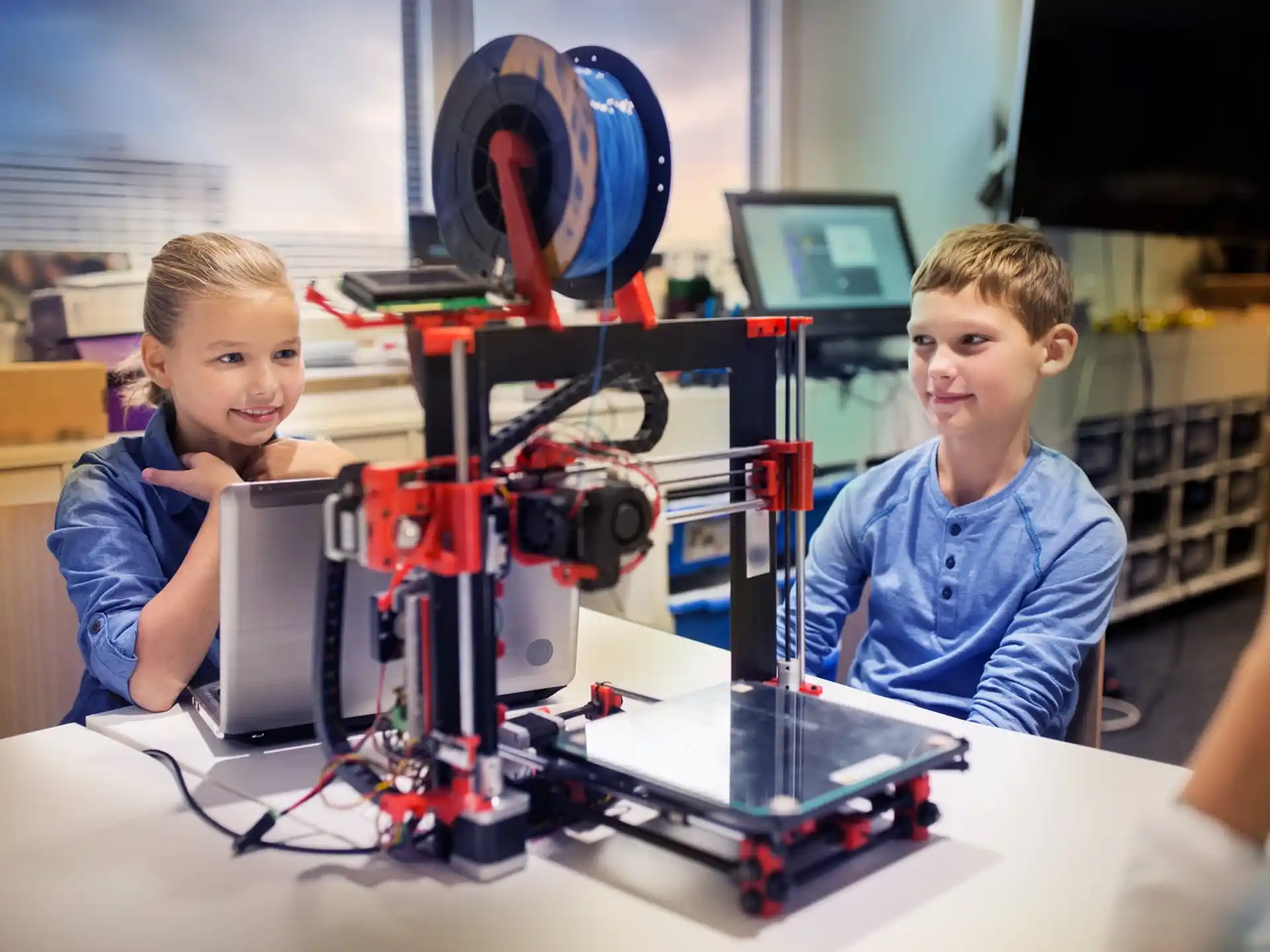 a couple of kids sitting at a table with a computer and a large projector