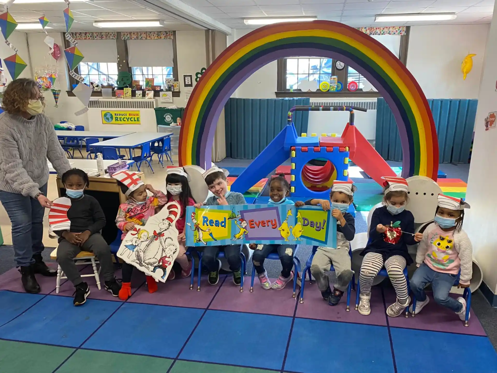 a group of children sitting on a mat in a room with a person standing and a person standing