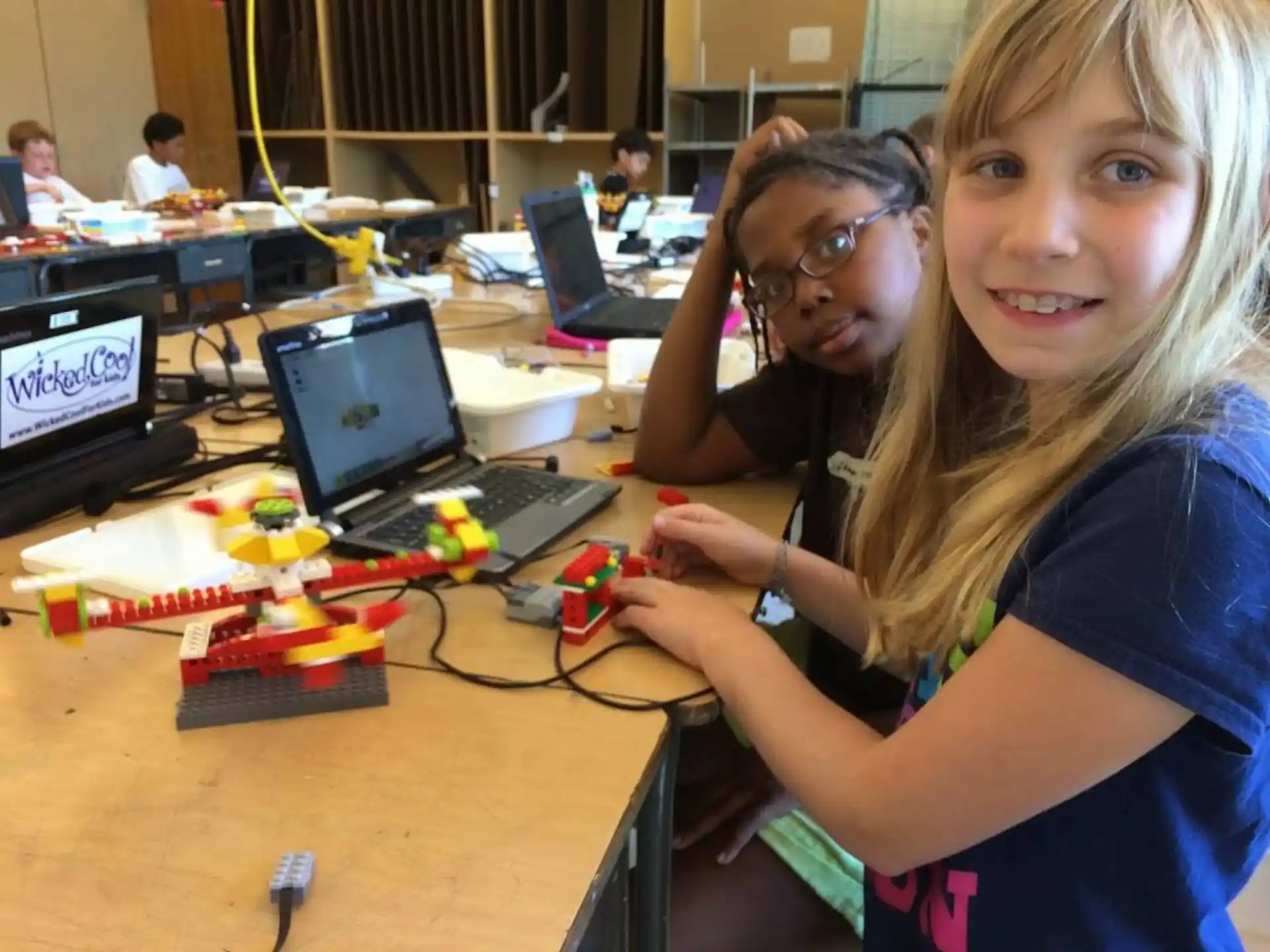 a couple of girls sitting at a table with computers and other people