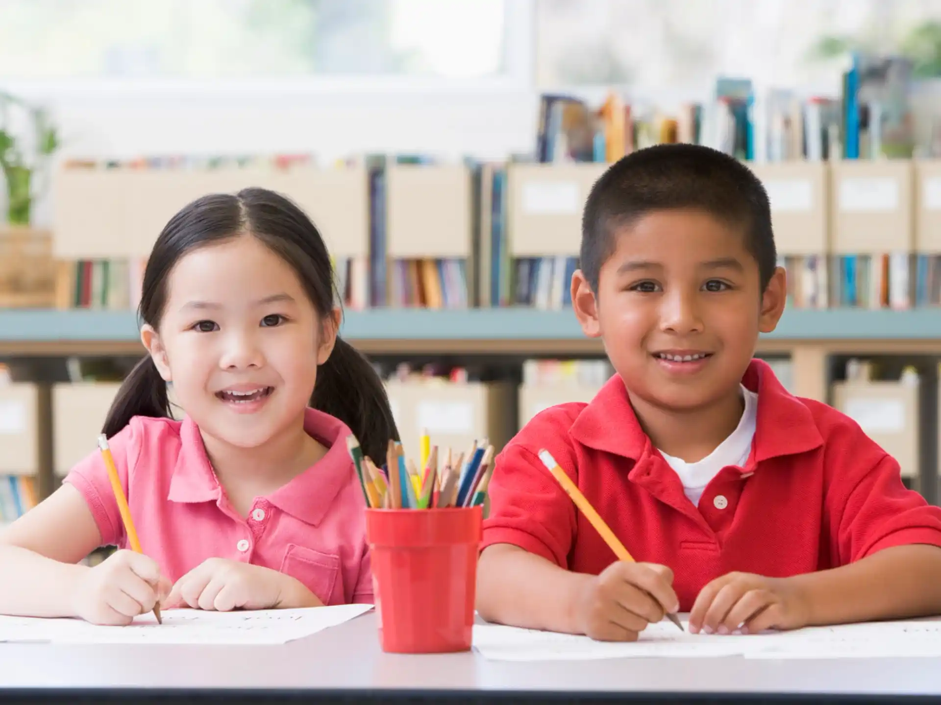 a couple of children sitting at a table with pencils and a cup